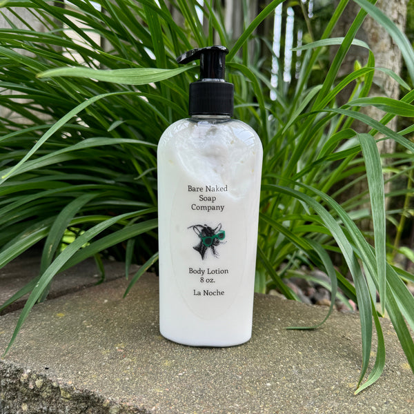 A pump bottle of body lotion with a label that reads 'Bare Naked Soap Company, Body Lotion 8 oz, La Noche'. The bottle is placed in front of a green leafy background.