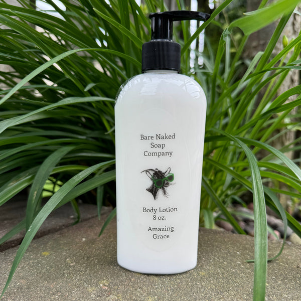 A pump bottle of Bare Naked body lotion labeled 'Amazing Grace' placed in front of green foliage.