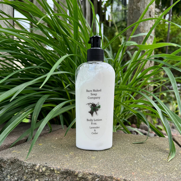 A pump bottle of body lotion labeled 'Bare Naked Soap Company Body Lotion 8 oz. Lavender & Cedar' placed on a stone surface with green plants in the background.