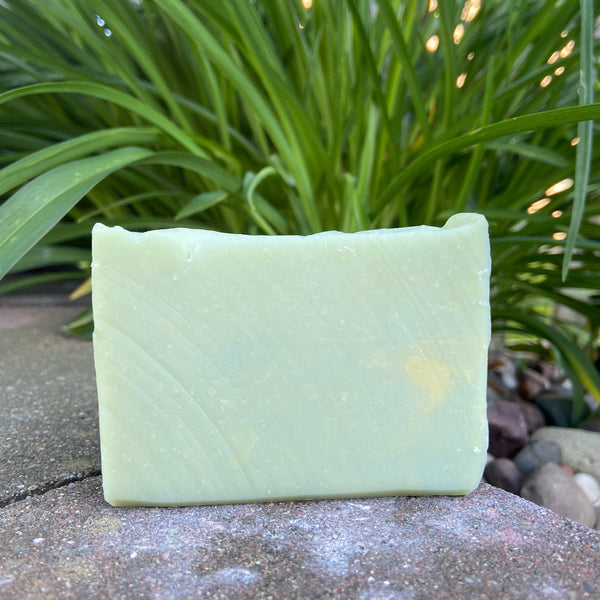 A bar of goat milk soap with a pale green hue, displayed outdoors with green foliage in the background.