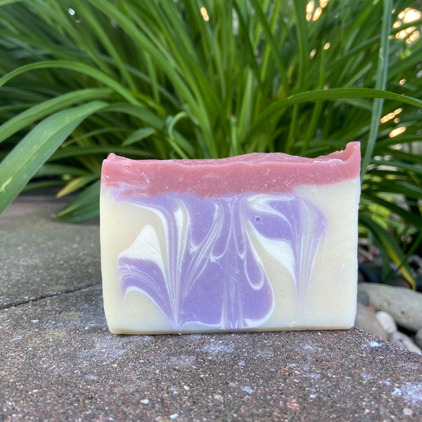 A bar of Cranberry & Pomegranate Goat Milk Soap with a swirled pattern in shades of cream, pink, and purple, placed on a surface with green foliage in the background.
