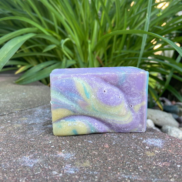 A bar of Energy Goat Milk Soap with a colorful marbled design featuring purple, blue, green, and yellow hues, placed on a stone surface with green foliage in the background.