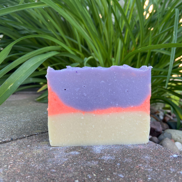 A bar of soap with layered colors of purple and orange, with a background of green foliage.