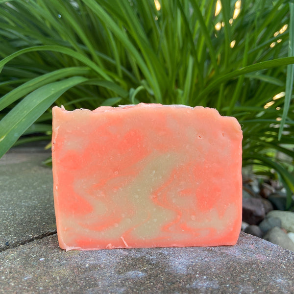 A bar of lavender chamomile soap with a pale orange to yellow gradient, placed outdoors with greenery in the background.
