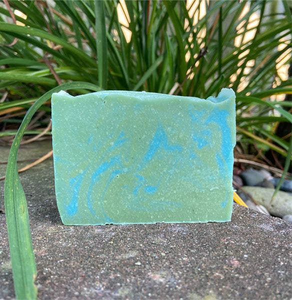 A bar of Lemongrass & Eucalyptus Goat Milk Soap with a green and blue marbled design, placed outdoors with foliage in the background.