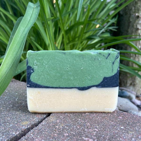 A bar of goat milk soap with a layered design featuring bands of green and blue, set on a natural stone surface with foliage in the background.
