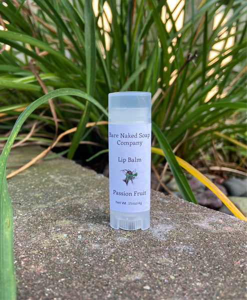 A stick of lip balm labeled 'Passion Fruit' from Pure Naked Soap Company, placed on a stone surface with grass in the background.
