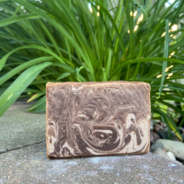 A bar of Sandalwood Vanilla Goat Milk Soap placed on a stone surface with green plants in the background.